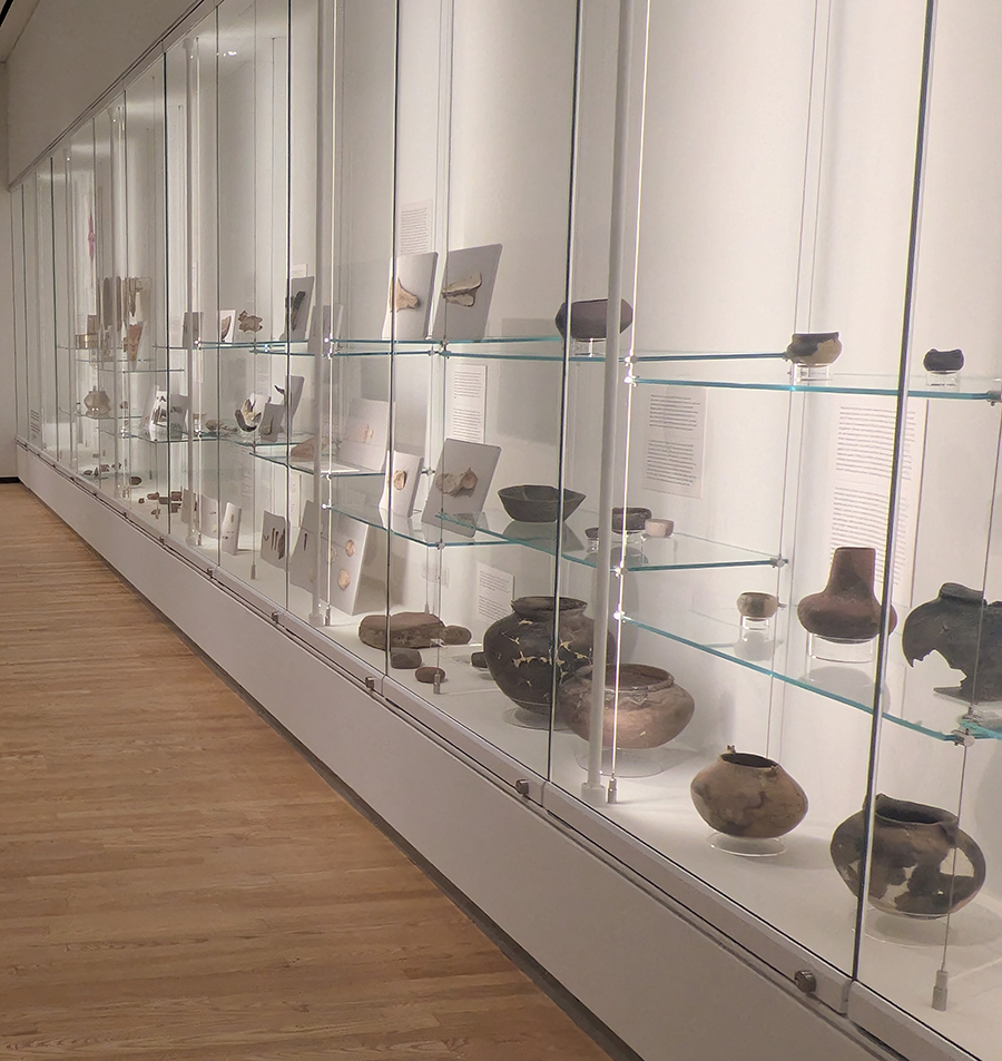 An arrangement of artifacts on glass shelves with a white background in an exhibit behind large glass windows
