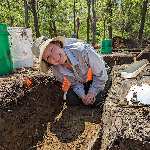 A woman with a sun hat kneeling in an excavation unit