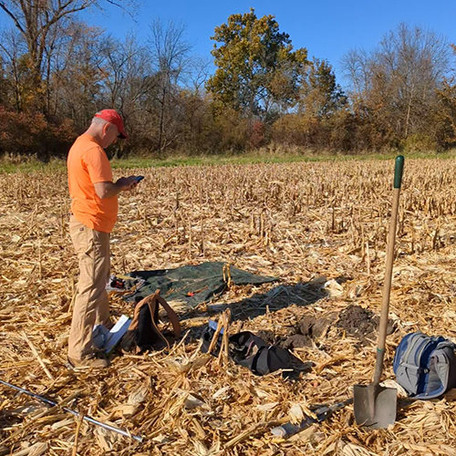 A man in an orange shirt takes notes while digging in a corn field