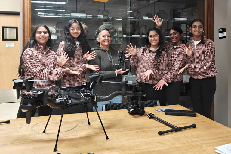 Five middle school girls in matching plaid shirts pose with a woman, standing behind a table with a drone set on top of it