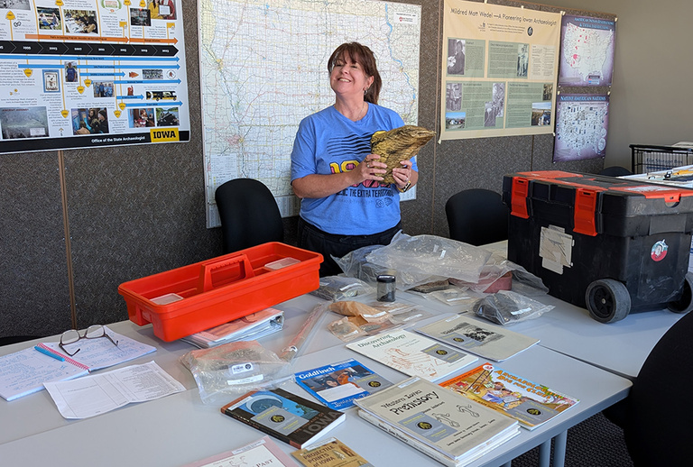 A woman smiles while holding a replica mammoth tooth, surrounded by teaching materials and books