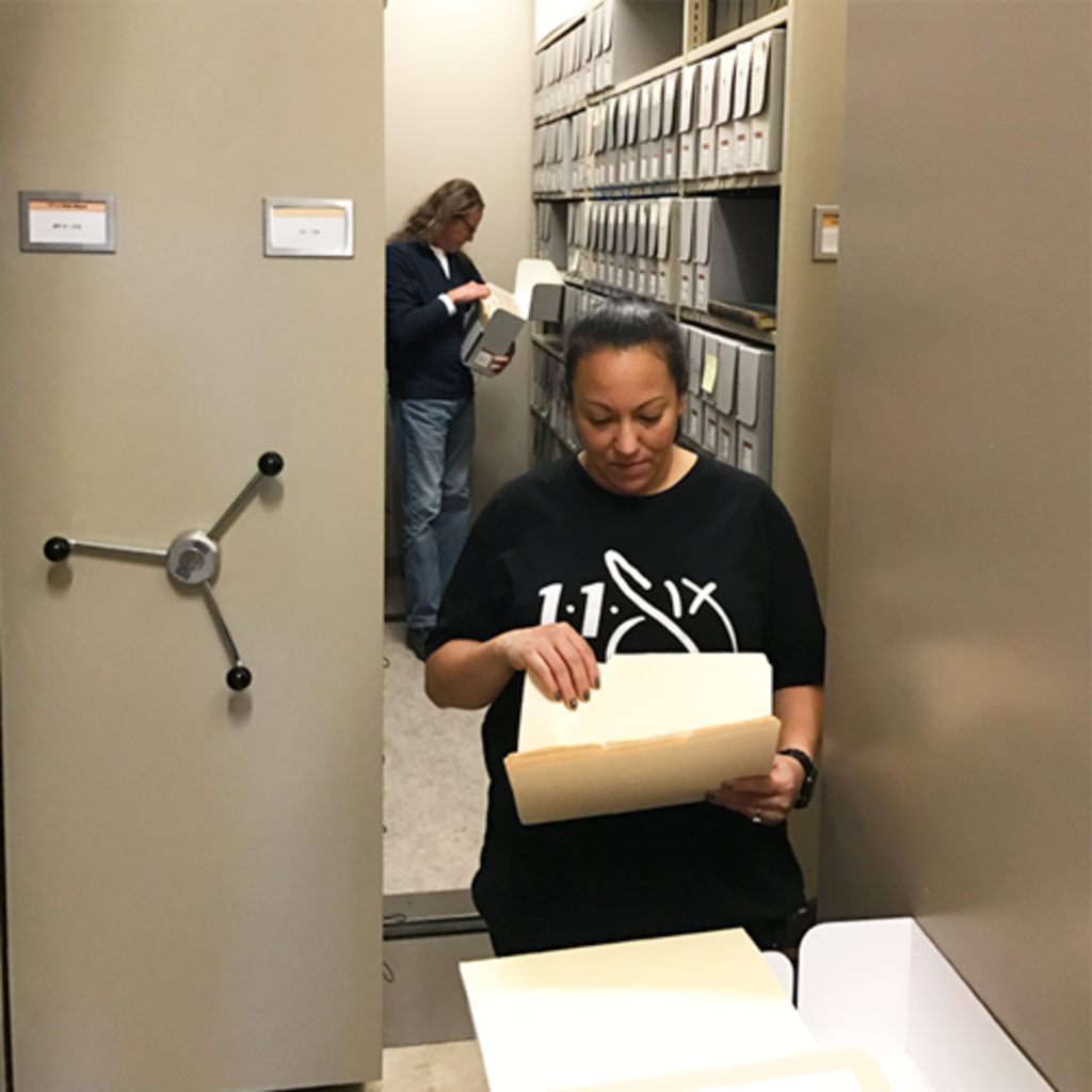 A women looks through file folders in an archives repository; a man is in the back in an aisle