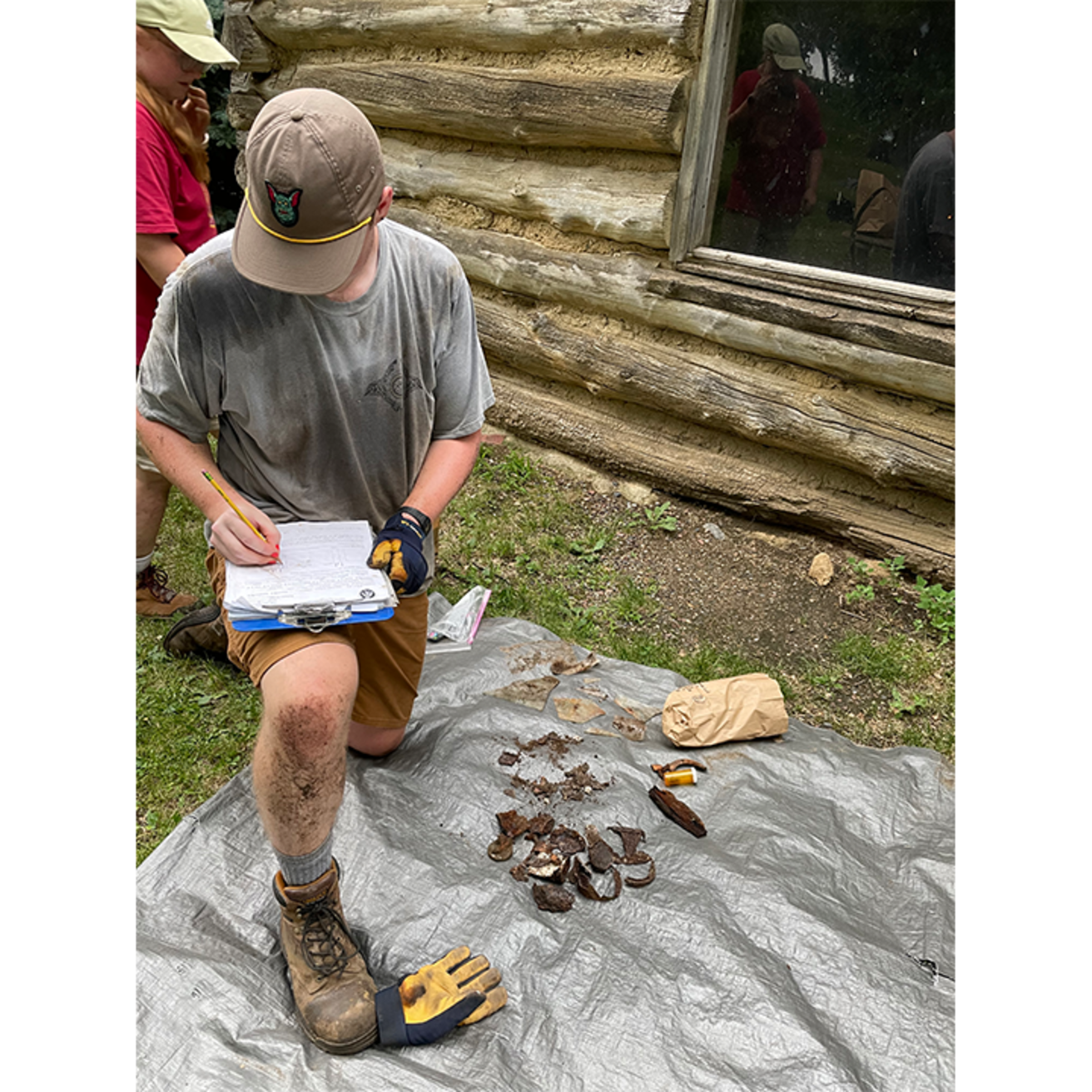 A student kneels on a tarp and takes notes about artifacts
