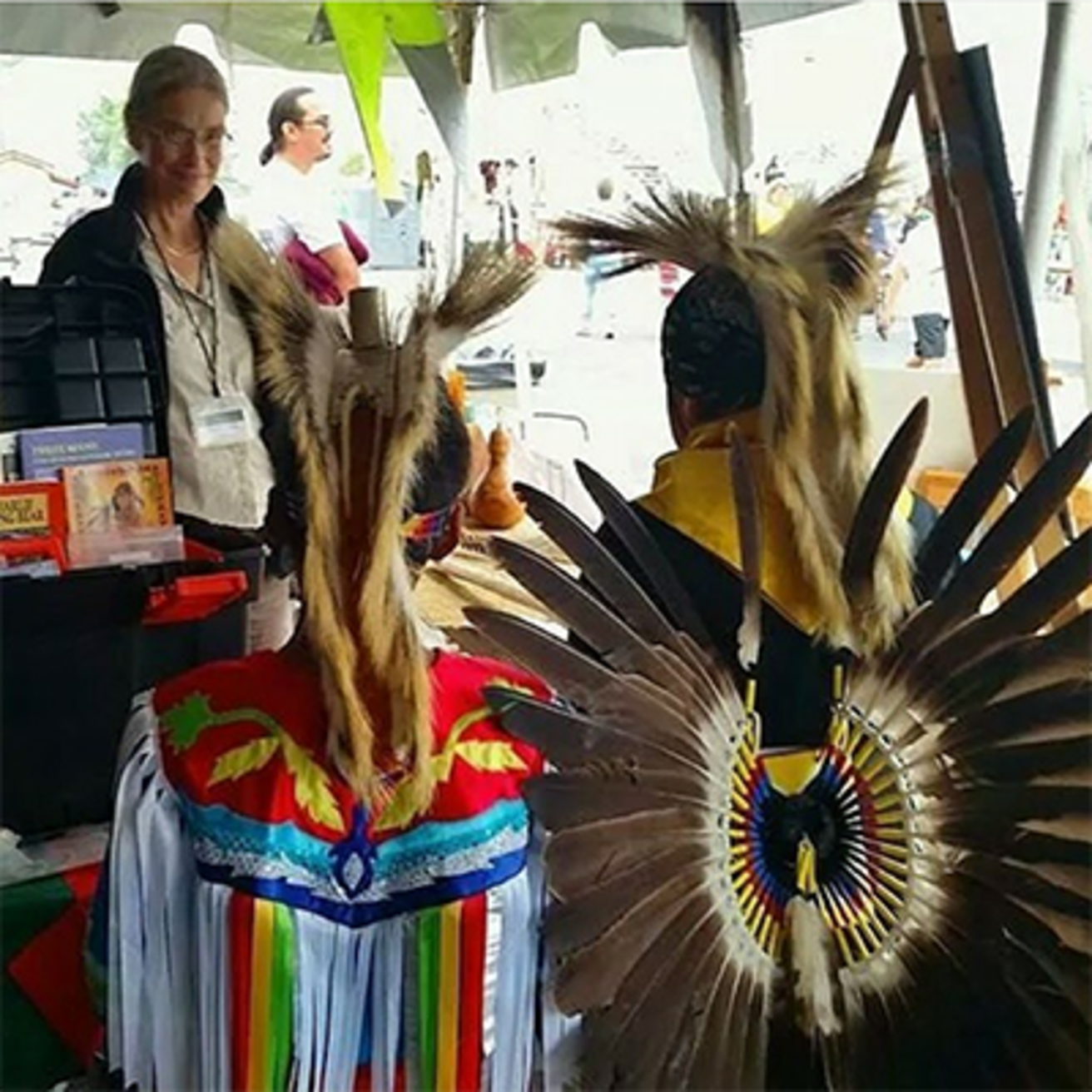 Two boys in regalia have their backs facing the camera. A lady on the others side of a table smiles at them