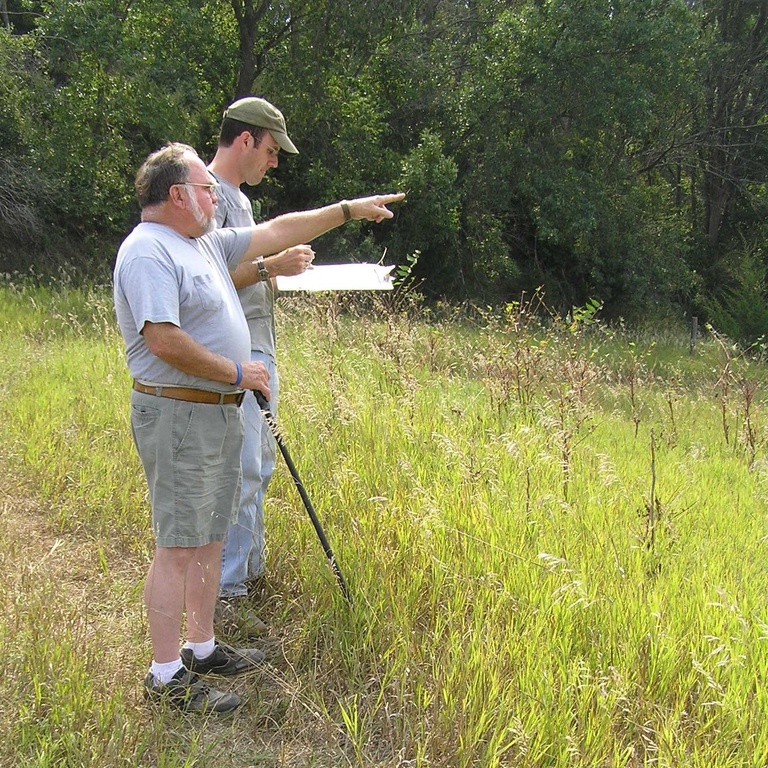 An archaeologist stands in a field with a clipboard, recording a site at the direction of a landowner.