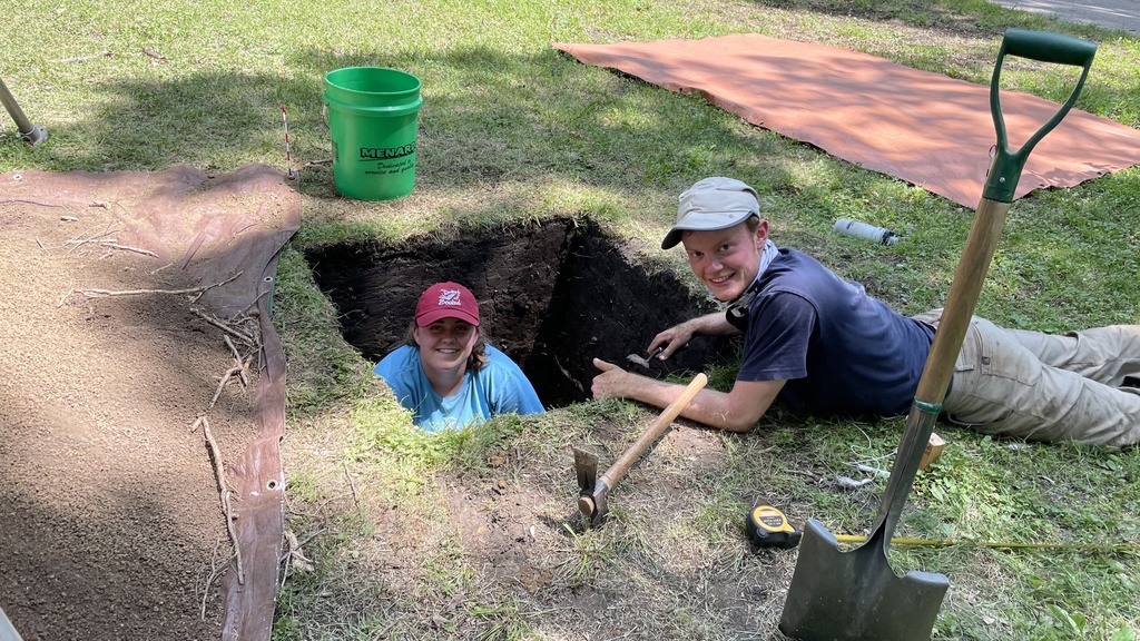 One student crouched in an excavation unit, one laying on his stomach next to the unit
