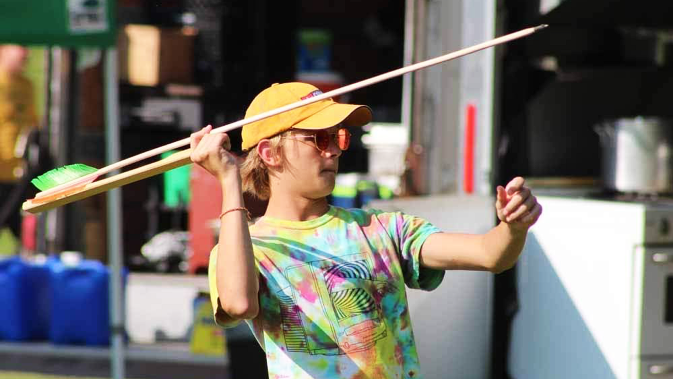 A boy in a tie dye shirt and yellow hat draws back a spear on an atlatl