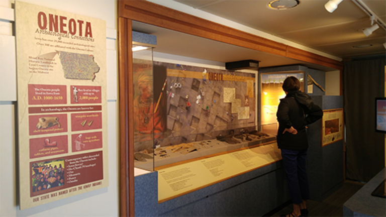 A women stands in front of a museum display that says "Oneota Archaeological Connections"